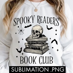Peut inclure: Sweat-shirt blanc avec le texte "Spooky Readers Book Club" et une illustration en noir et blanc d'un crâne sur une pile de livres avec des chauves-souris et des araignées.
