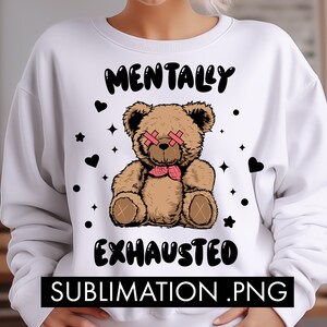 May include: White sweatshirt with a brown teddy bear wearing pink eye patches and a pink bow tie. The text "Mentally Exhausted" is printed in black on the sweatshirt.