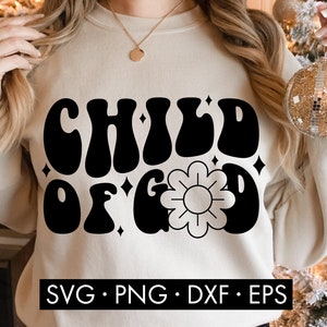 May include: A beige sweatshirt with the text "CHILD OF GOD" in black, retro-style lettering. A stylized flower is centered below the text. The sweatshirt is being worn by a person.