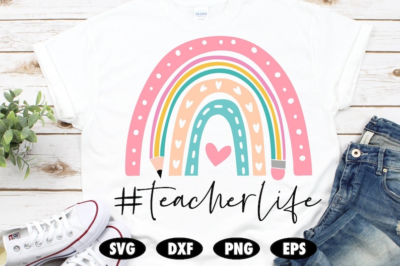 K&ouml;nnte beinhalten: Ein wei&szlig;es T-Shirt mit einem Regenbogen-Design mit einem Herzen und Bleistiften. Der Text "#teacherlife" ist unter dem Regenbogen gedruckt.