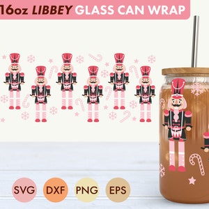 May include: A 16oz glass can wrap featuring a pink and red nutcracker design. The design includes candy canes, snowflakes, and stars. The wrap is shown on a glass can with a brown liquid inside. The image also includes the text "SVG, DXF, PNG, EPS".