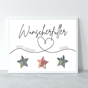 May include: A white framed print with the text "Wunschersch&uuml;ller" in black cursive font. Below the text is a heart shape with a line extending to the left with the text "HAPPY BIRTHDAY" and a line extending to the right with the text "THOMAS". Three stars made of folded money are placed below the lines.