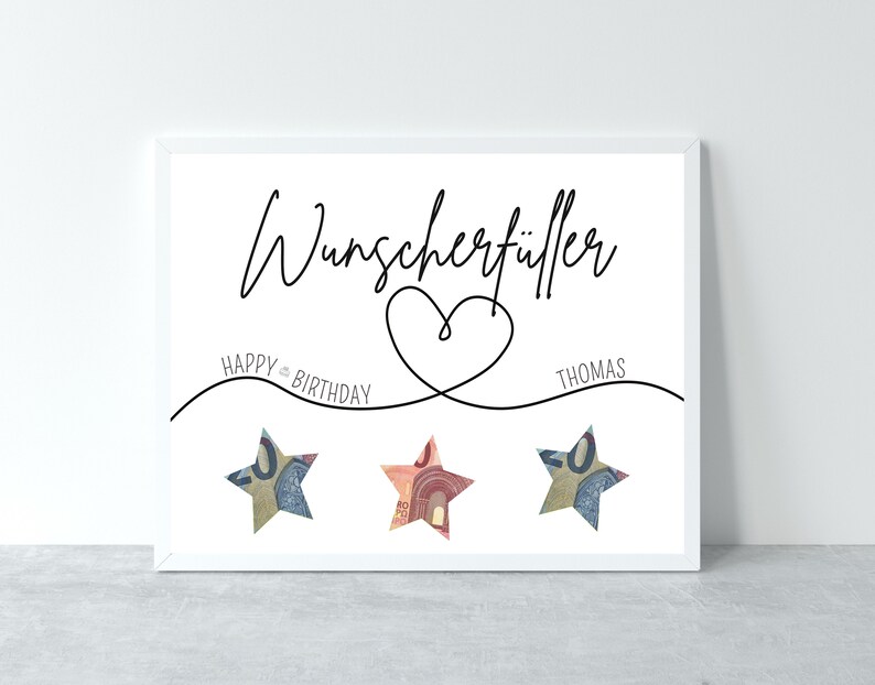 May include: A white framed print with the text "Wunschersch&uuml;ller" in black cursive font. Below the text is a heart shape with a line extending to the left with the text "HAPPY BIRTHDAY" and a line extending to the right with the text "THOMAS". Three stars made of folded money are placed below the lines.