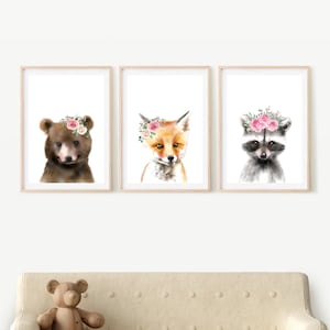May include: Three framed watercolor paintings of woodland animals wearing flower crowns. The animals are a bear, a fox, and a raccoon. The paintings are on a white background and are framed in natural wood.