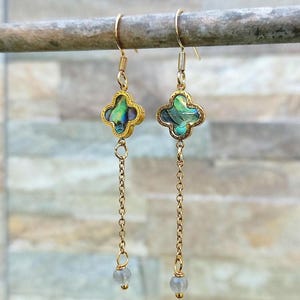Può includere: Un paio di orecchini pendenti color oro. Ogni orecchino presenta un ciondolo a forma di quadrifoglio con intarsi iridescenti di conchiglia di abalone verde e blu. Una delicata catena dorata pende sotto, terminando con una piccola perla grigio chiaro.