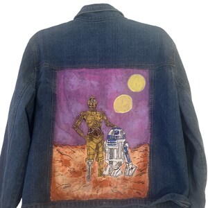 May include: A denim jacket with a painted back panel featuring C-3PO and R2-D2 standing on a desert planet with two suns in the sky. The painting is done in shades of purple, yellow, orange, and brown.