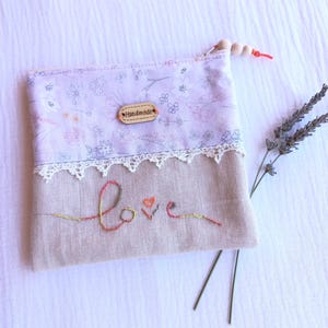 Peut inclure: Une petite pochette faite à la main avec un dessus à motifs floraux et un bas beige. Le mot "love" est brodé avec du fil coloré. Une étiquette en bois indique "Handmade". Une brindille de lavande repose à côté de la pochette.