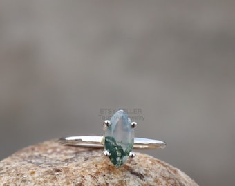 May include: A silver ring with a marquise-shaped green moss agate gemstone. The ring is set on a rock.