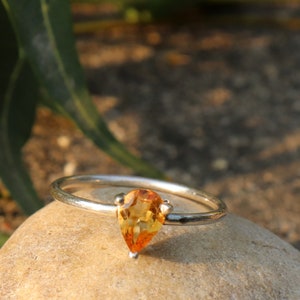 May include: A silver ring with a pear-shaped, orange gemstone. The ring is set on a light-colored stone, with green foliage in the background. The gemstone has a vibrant, warm color, and the ring band is thin and delicate.