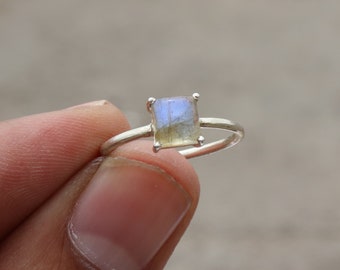 May include: A silver ring with a square-shaped labradorite gemstone. The gemstone has a light blue and gray color with iridescent flashes.