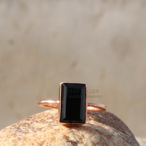 May include: A black gemstone ring with a rectangular cut stone set in a rose gold band. The ring is sitting on a rock.
