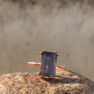 Puede incluir: Un anillo de oro rosa con una piedra rectangular gris oscuro. El anillo está colocado sobre una roca marrón.