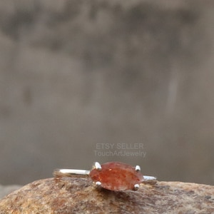 May include: A silver ring with a reddish-orange sunstone gemstone. The ring is set on a brown rock.