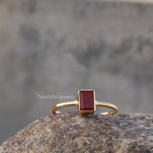 Op de afbeelding: Een gouden ring met een rechthoekige rode edelsteen. De ring ligt op een bruine steen.