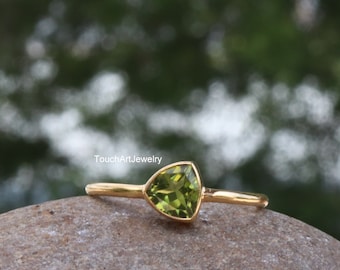May include: A gold ring with a triangular green gemstone. The ring is on a brown rock with a blurred green background.