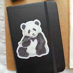 Op de afbeelding: Een zwart leren notitieboek met een elastische sluiting en een panda-sticker. De panda is zwart en wit en houdt bamboe vast. Een inktkussen en houten letterblokken staan op de achtergrond.