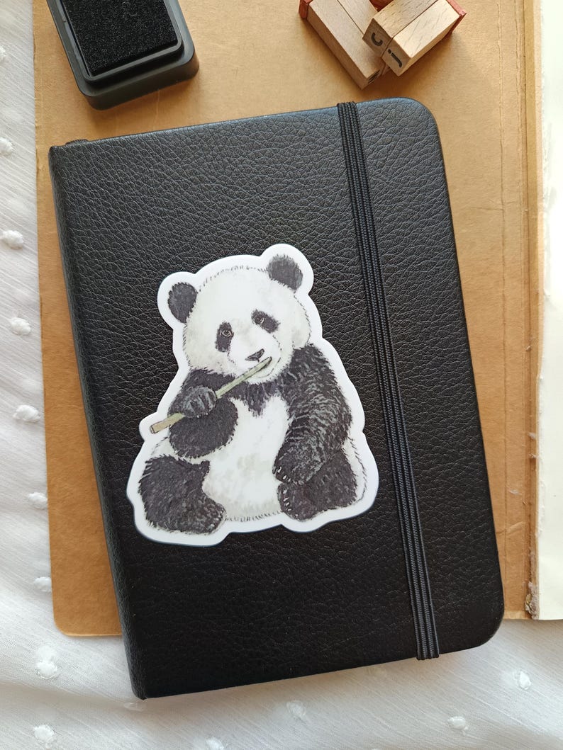 Op de afbeelding: Een zwart leren notitieboek met een elastische sluiting en een panda-sticker. De panda is zwart en wit en houdt bamboe vast. Een inktkussen en houten letterblokken staan op de achtergrond.