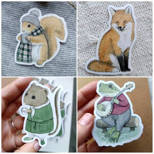 Op de afbeelding: Vier grillige stickers met bosdieren. Bevat een eekhoorn met een mok, een vos, een muis in een jas en een kikker die banjo speelt. Elke sticker heeft een witte rand en staat op een neutrale achtergrond.