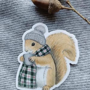 Op de afbeelding: Een sticker met een cartoon eekhoorn met een grijze gebreide muts en een groen-zwarte geruite sjaal. De eekhoorn houdt een grijze mok vast. De eekhoorn heeft een pluizige bruine staart.