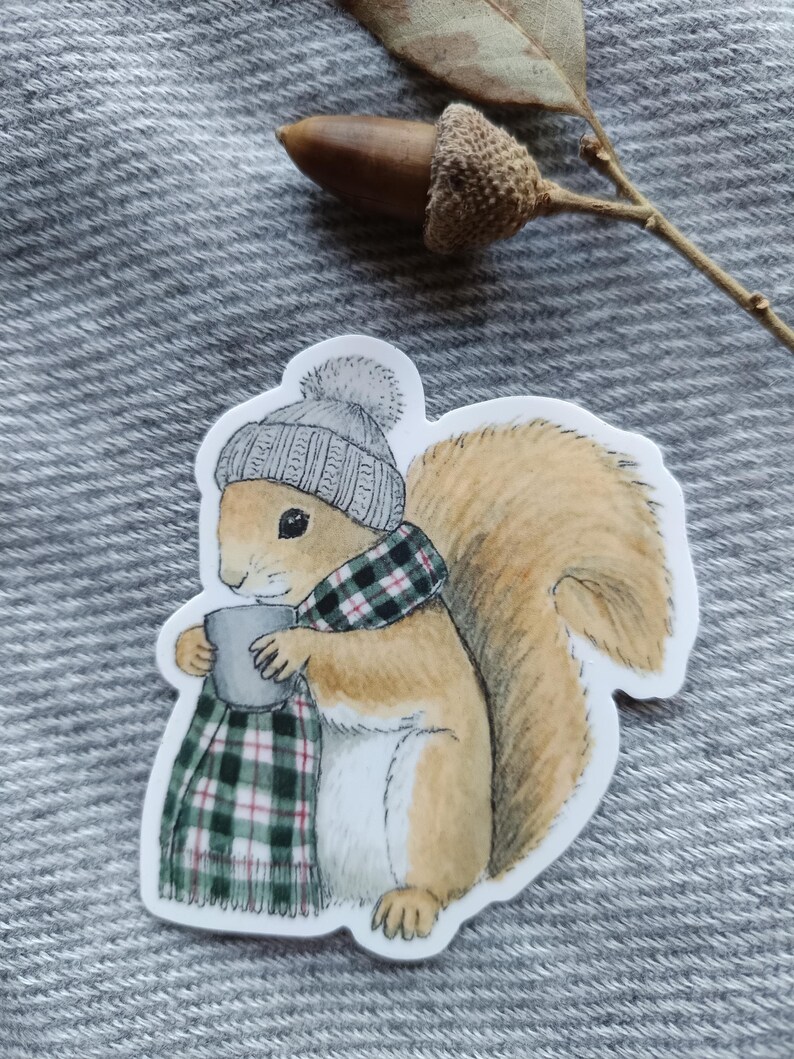 Op de afbeelding: Een sticker met een cartoon eekhoorn met een grijze gebreide muts en een groen-zwarte geruite sjaal. De eekhoorn houdt een grijze mok vast. De eekhoorn heeft een pluizige bruine staart.