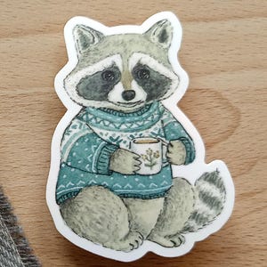 Op de afbeelding: Een sticker met een wasbeer die een teal trui draagt en een mok koffie vasthoudt. De wasbeer heeft een grijs en zwart gezicht en een gestreepte staart. De mok heeft een bloemenontwerp.