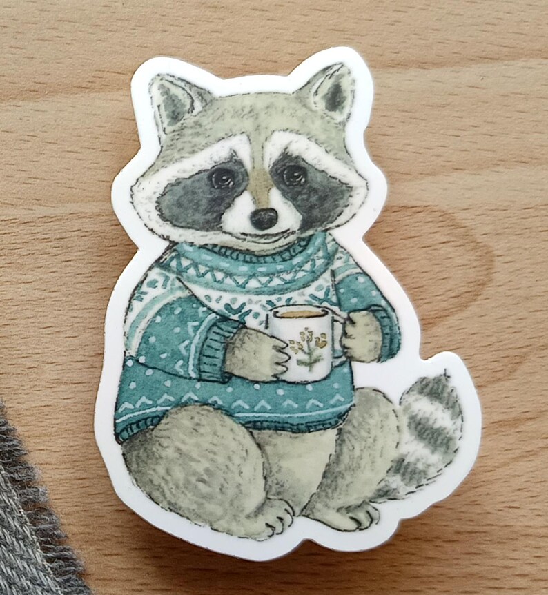 Op de afbeelding: Een sticker met een wasbeer die een teal trui draagt en een mok koffie vasthoudt. De wasbeer heeft een grijs en zwart gezicht en een gestreepte staart. De mok heeft een bloemenontwerp.