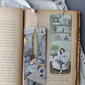 Può includere: Due segnalibri illustrati con nappe argentate su un libro aperto. Uno raffigura una donna a un tavolo con un laptop e un caffè, l'altro una donna che legge su una sedia. I segnalibri hanno un tema accogliente e libresco.