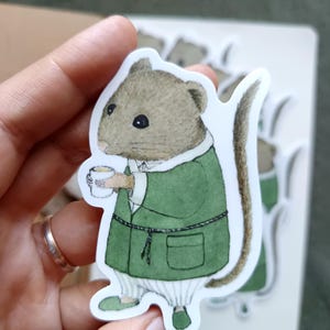 Op de afbeelding: Een sticker met een charmant muispersonage in een groene jurk, met een kopje in de hand. De muis heeft een bruine kop en een lange staart. De sticker staat op een witte achtergrond, met andere stickers zichtbaar.