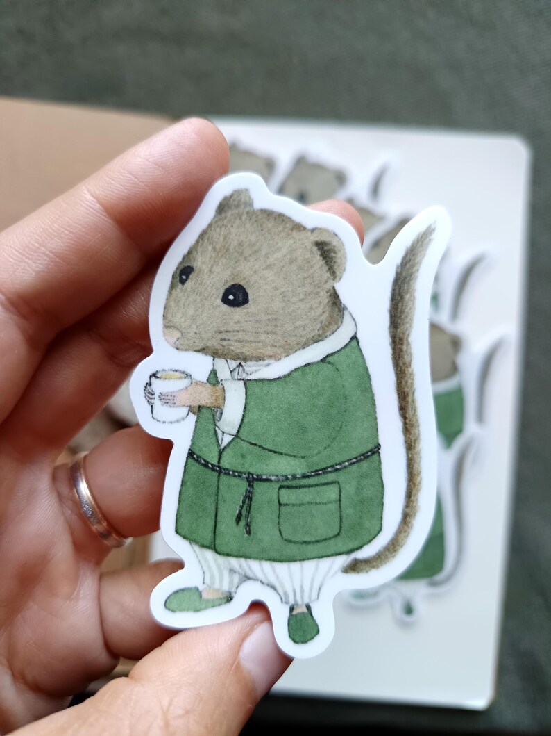 Op de afbeelding: Een sticker met een charmant muispersonage in een groene jurk, met een kopje in de hand. De muis heeft een bruine kop en een lange staart. De sticker staat op een witte achtergrond, met andere stickers zichtbaar.