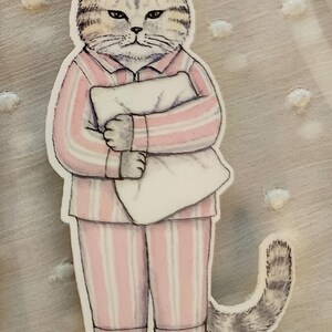 Op de afbeelding: Een sticker met een chagrijnige kat in een roze en wit gestreepte pyjama, die een wit kussen vasthoudt. De kat heeft grijs en bruin gestreepte vacht en een bijpassende gestreepte staart. Een grillige illustratie.