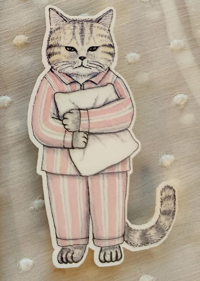Op de afbeelding: Een sticker met een chagrijnige kat in een roze en wit gestreepte pyjama, die een wit kussen vasthoudt. De kat heeft grijs en bruin gestreepte vacht en een bijpassende gestreepte staart. Een grillige illustratie.