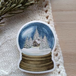 May include: A sticker depicting a snow globe. The design shows a winter scene with a house and snow-covered trees inside a blue globe with white snowflakes. The base of the snow globe is brown.
