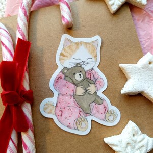 Op de afbeelding: Een illustratie van een kat in een roze pyjama die een teddybeer knuffelt, omringd door zuurstokken en stervormige koekjes. De kat heeft oranje en witte vacht. Een rood fluwelen lint siert de zuurstokken. De sc&egrave;ne roept een gezellige, feestelijke sfeer op.