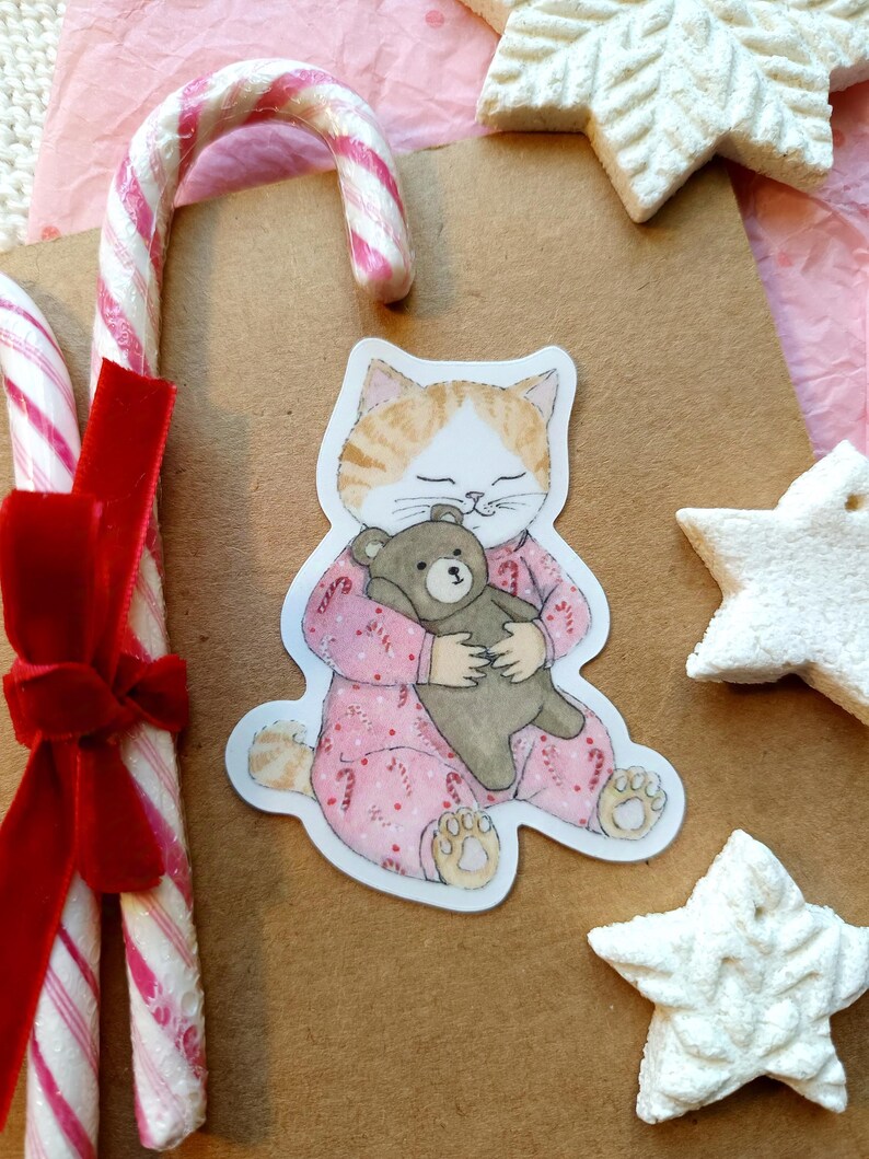 Op de afbeelding: Een illustratie van een kat in een roze pyjama die een teddybeer knuffelt, omringd door zuurstokken en stervormige koekjes. De kat heeft oranje en witte vacht. Een rood fluwelen lint siert de zuurstokken. De sc&egrave;ne roept een gezellige, feestelijke sfeer op.