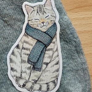 Op de afbeelding: Een sticker met een tabby kat met een blauwe sjaal. De kat heeft de ogen gesloten en een serene uitdrukking. De sticker heeft een witte rand en ligt op een grijze ondergrond.