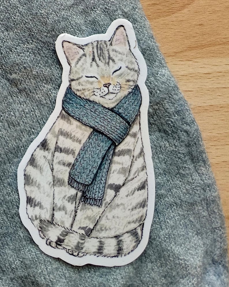 Op de afbeelding: Een sticker met een tabby kat met een blauwe sjaal. De kat heeft de ogen gesloten en een serene uitdrukking. De sticker heeft een witte rand en ligt op een grijze ondergrond.