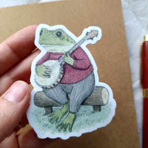 Op de afbeelding: Een sticker met een groene kikker die banjo speelt, zittend op een houten bank. De kikker draagt een rode trui en een grijze broek. De sticker heeft een witte rand.