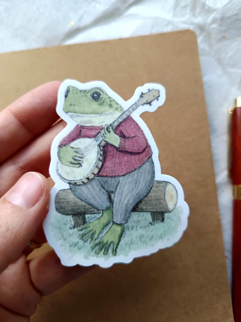 Op de afbeelding: Een sticker met een groene kikker die banjo speelt, zittend op een houten bank. De kikker draagt een rode trui en een grijze broek. De sticker heeft een witte rand.