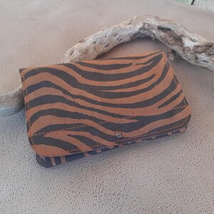 May include: A brown and black zebra print wallet with a snap closure. The wallet is made of a textured material and has a stitched border. The wallet is closed and resting on a neutral surface.