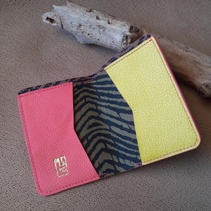 May include: Open, colourful wallet with a pink exterior, a yellow interior, and a zebra print accent. The wallet has a gold logo and is stitched with pink thread. The background is a textured brown surface.