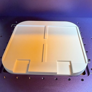 May include: A white, flat, rectangular 3D printed object with rounded corners, featuring a central vertical divider and two symmetrical recessed square cutouts along the front edge, resting on a purple textured surface with a grid of small holes.