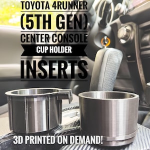 以下が含まれることがあります： トヨタ 4ランナー（5代目）センターコンソールの3Dプリントされた黒いカップホルダーインサート2個。画像の下部に「3D PRINTED ON DEMAND!」のテキストがあります。