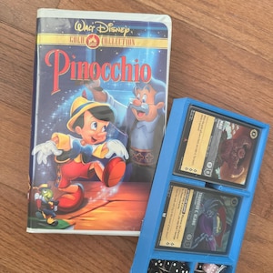 May include: A blue plastic card game organizer with six compartments. The organizer has two cards in it, one with a purple creature and the other with a red creature. There are also six black dice in the organizer. The organizer is sitting on a wooden surface next to a VHS tape of the Disney movie Pinocchio.