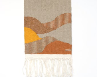 May include: A woven wall hanging with a beige background and abstract shapes in shades of brown, orange, and yellow. The hanging has a white fringe and is attached to a wooden dowel with a white cord.