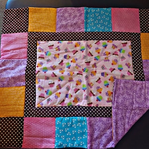 May include: A patchwork quilt with a brown, white, and pink polka dot border. The center of the quilt is a pink fabric with a cupcake pattern. The quilt is made up of various colors and patterns, including purple, blue, and yellow.