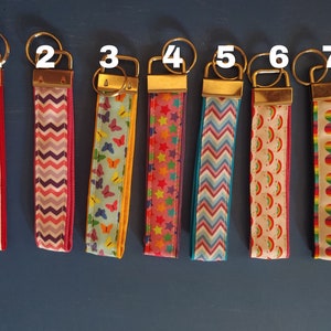 May include: Seven keychains with gold-colored hardware and patterned fabric straps. Designs include owls, chevrons, butterflies, stars, and rainbows. The keychains are arranged on a blue surface.