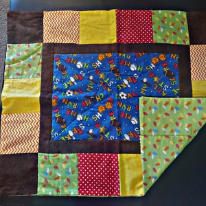Op de afbeelding: Een patchwork quilt met een sportthema. Het middenstuk heeft een blauwe stof met sportartikelen en tekst. De rand bevat bruine, gele, groene en rood gekleurde vierkanten. De quilt is ontworpen voor een kind.