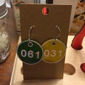 以下が含まれることがあります： 茶色のカードに展示された、丸いイヤリングのペア。片方は緑色で白い数字の「061」が、もう片方は黄色で白い「031」が描かれています。イヤリングにはシルバーのフックが付いています。