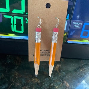 May include: Novelty earrings in the shape of pencils. The earrings have a yellow and orange body, a pink eraser, and a silver-coloured hook. The pencils are labelled "No. 2 HB".