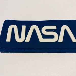 Może przedstawiać: Niebieska łatka z białym haftem z napisem "NASA".